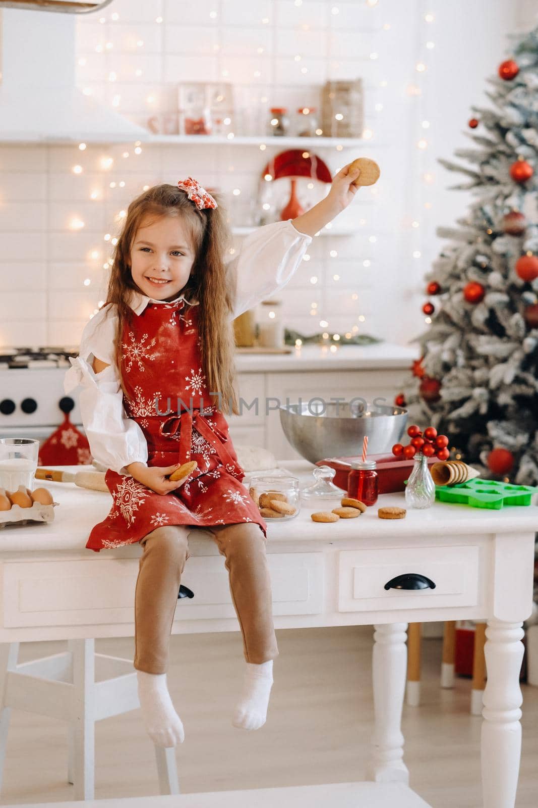 A happy little girl in the Christmas kitchen is sitting on the table with cookies in her hands by Lobachad