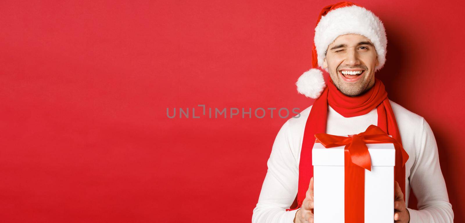 Concept of winter holidays, christmas and lifestyle. Handsome cheeky man in santa hat and scarf, holding present and smiling, winking at camera, standing over red background by Benzoix