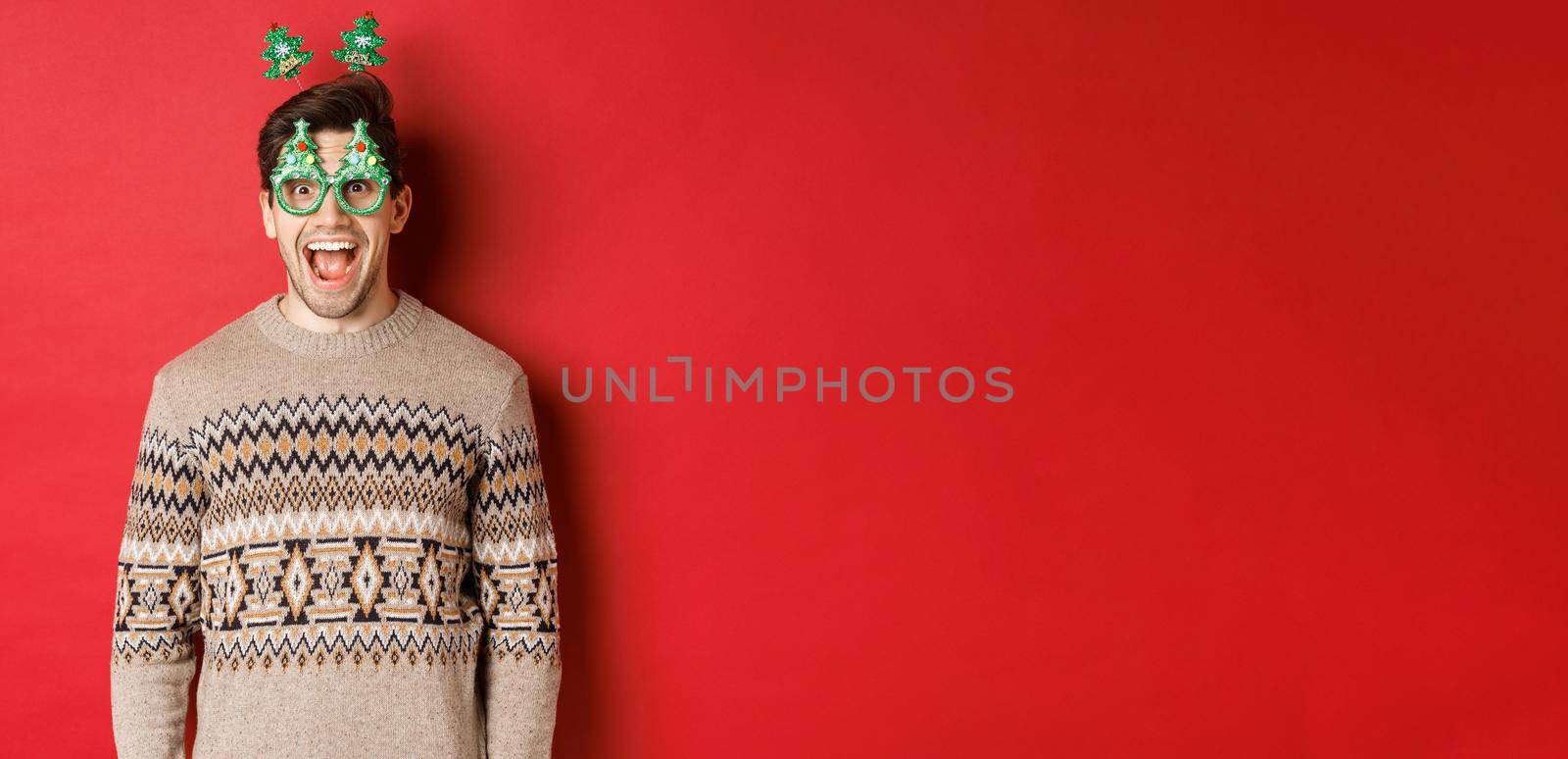 Portrait of surprised and excited handsome guy in party glasses and winter sweater, celebrating christmas and having fun, standing against red background.