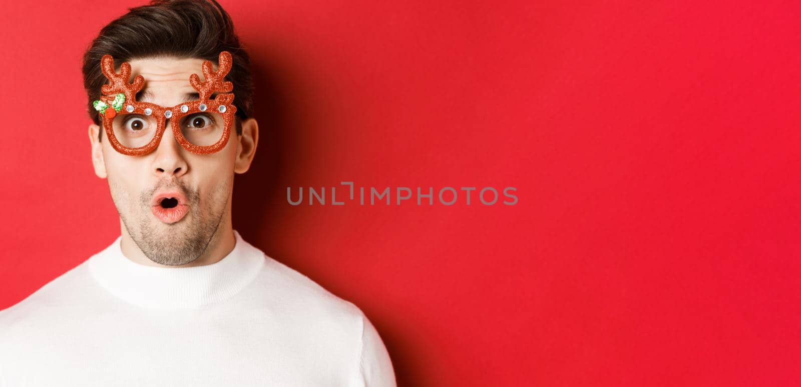Concept of winter holidays, christmas and celebration. Close-up of attractive guy with bristle, wearing new year party glasses and looking impressed at promo offer, standing over red background.