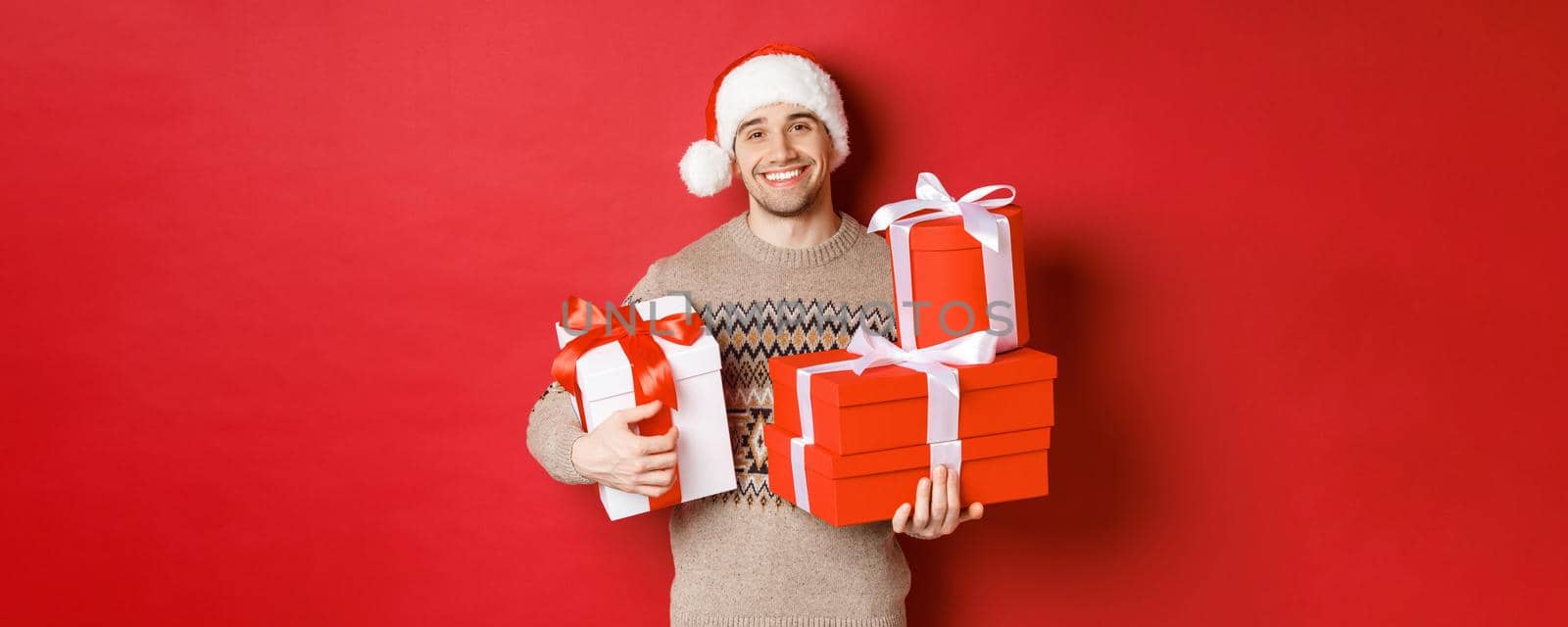 Concept of winter holidays, new year and celebration. Portrait of lovely smiling man prepared gifts for christmas, holding presents and looking at camera heartwarming, red background by Benzoix