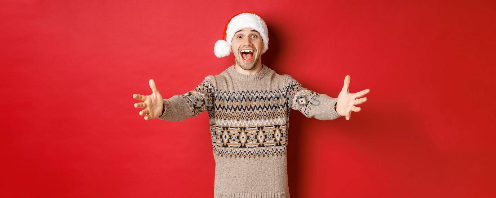 Portrait of happy, attractive man in christmas sweater and santa hat, reaching hands for gift, want to take something and smiling, standing over red background.