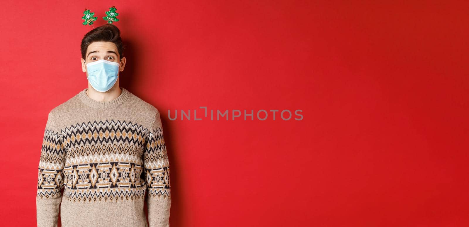 Concept of christmas, covid-19 and social distancing. Cheerful young man in medical mask and new year clothing, celebrating holidays during pandemic, standing over red background by Benzoix