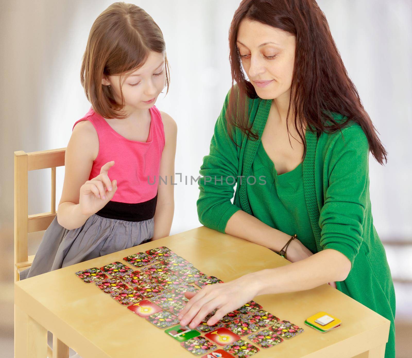 Pensive little girl and her teacher at the table laid out cards with pictures.In a room with a large semi-circular window.