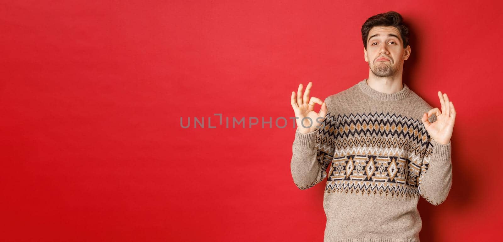 Impressed handsome man in christmas sweater, showing okay signs and praising good party, nodding in approval, standing pleased against red background.