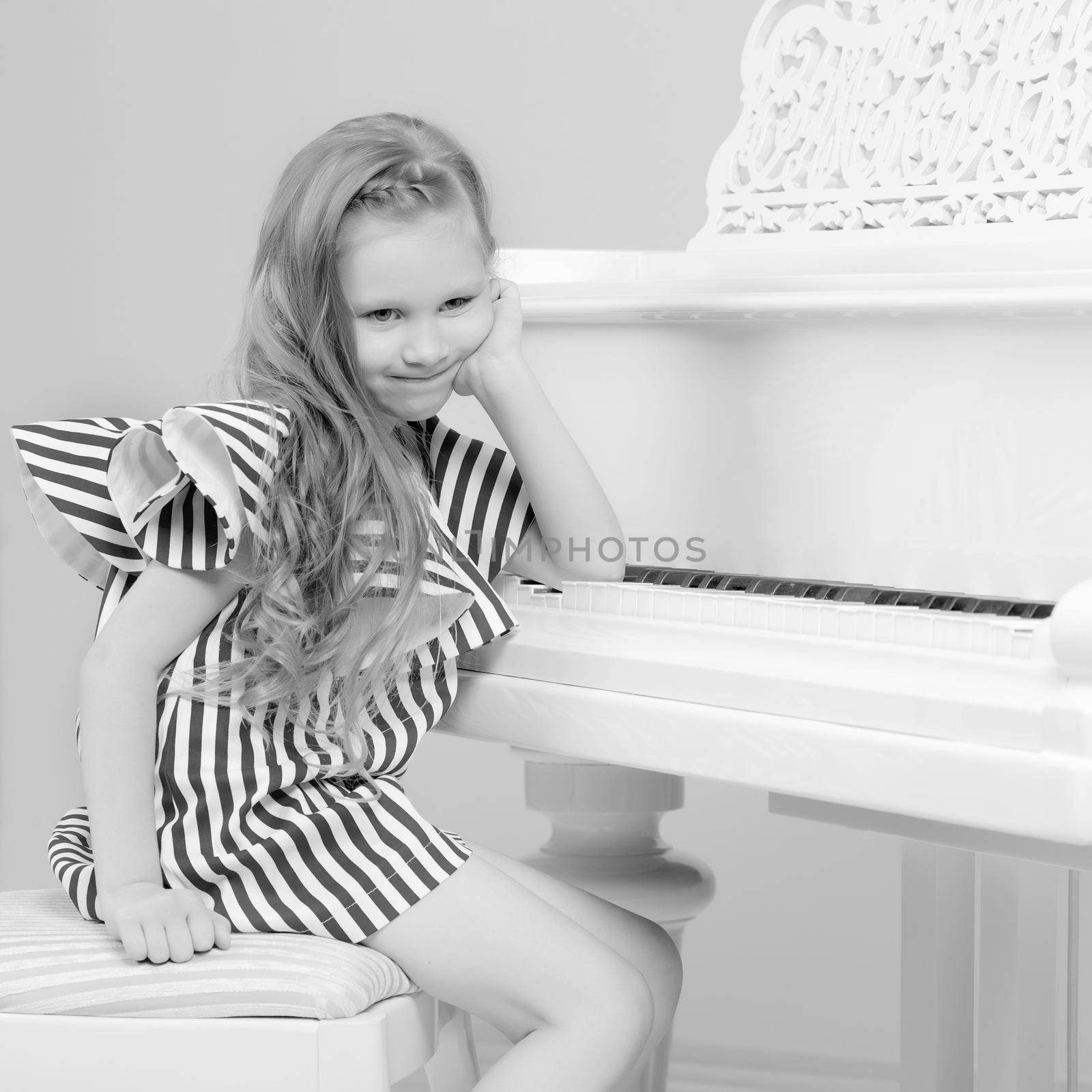 Adorable little blond girl in very short summer striped dress.She sits on a chair near a white piano.Black and white photography.