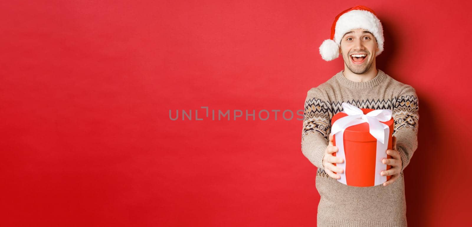 Image of happy man saying merry christmas and giving you a gift in box, smiling cheerful, celebrating winter holidays in santa hat and xmas sweater, standing over red background by Benzoix
