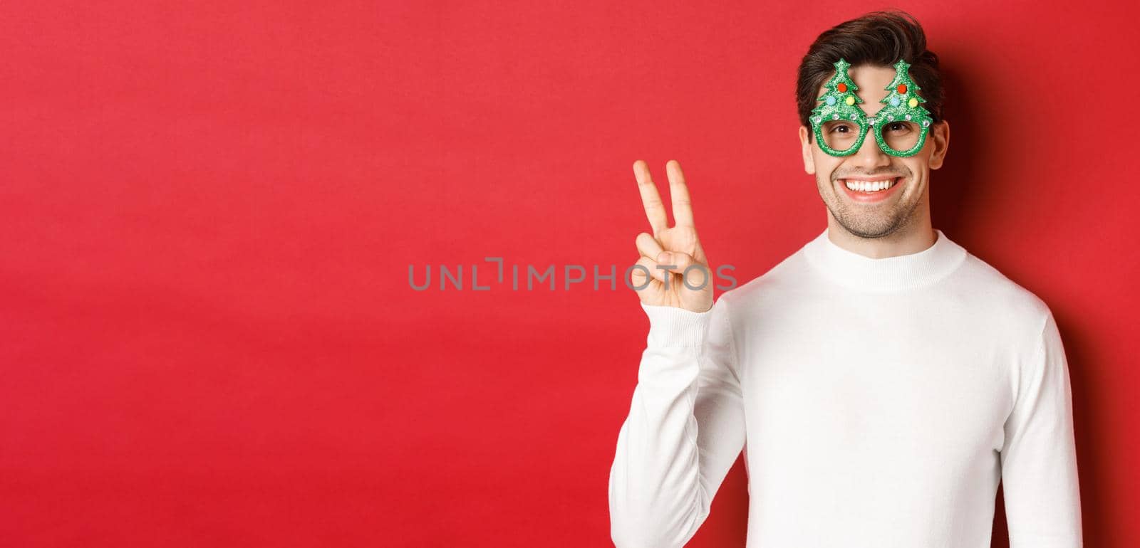 Image of handsome man in white sweater and party glasses, showing peace sign and smiling, wishing merry christmas, standing over red background by Benzoix