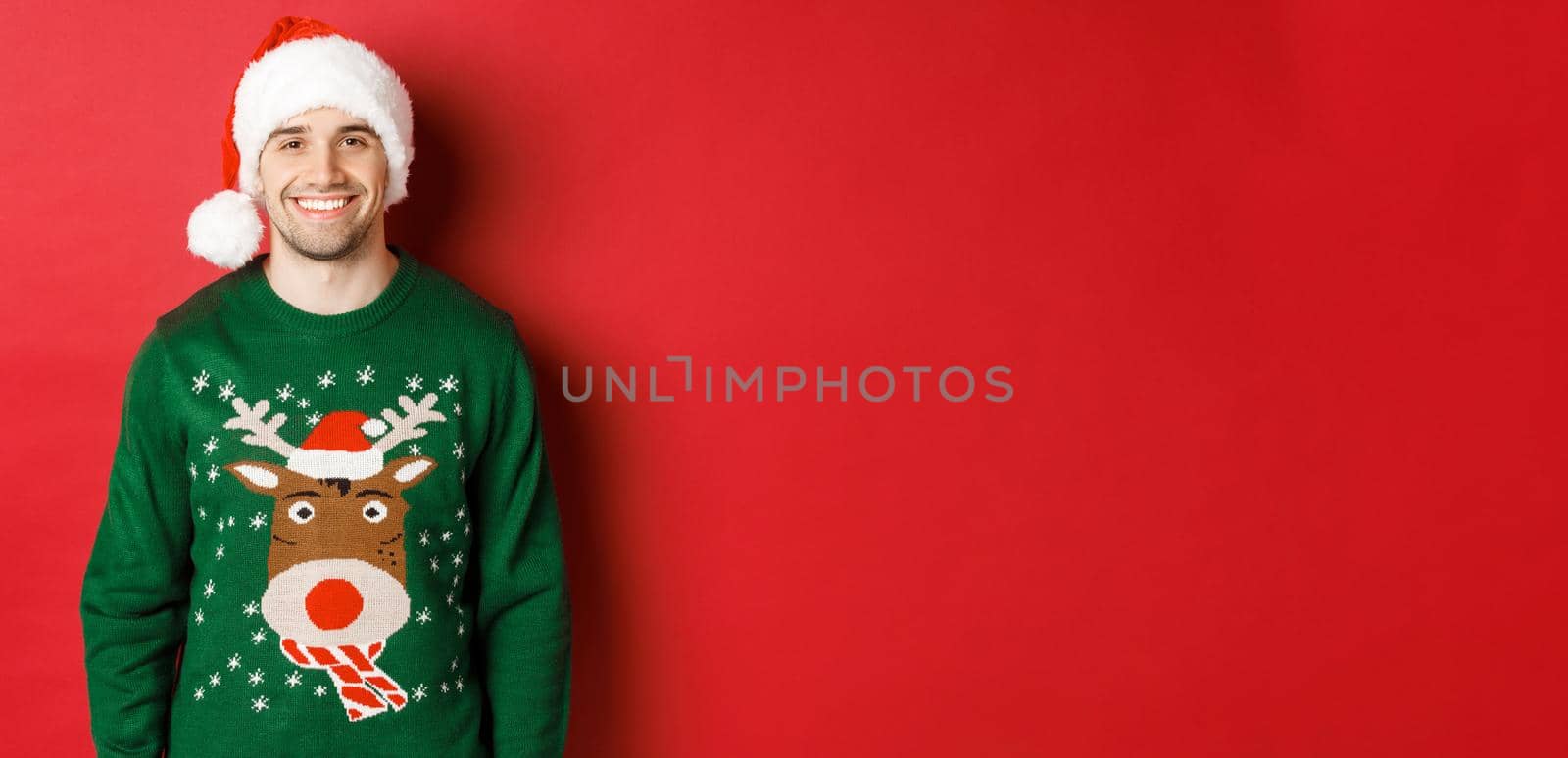 Concept of winter holidays, christmas and lifestyle. Handsome guy with bristle, wearing santa hat and green sweater, smiling joyful, celebrating new year, standing over red background by Benzoix