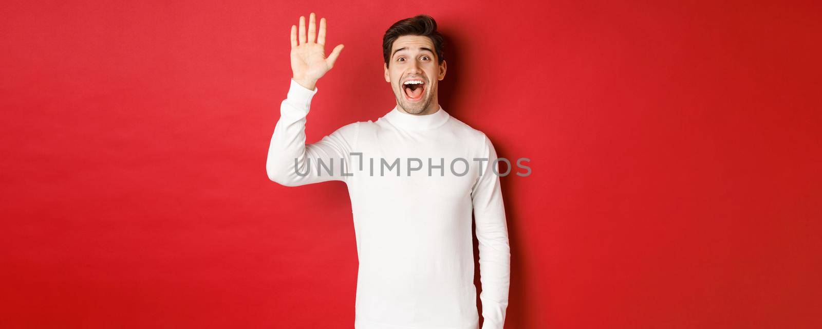 Image of happy and friendly young man saying hello, waving hand to greet someone, standing over red background.