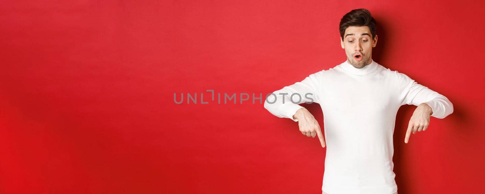 Portrait of surprised good-looking guy in white sweater, looking and pointing fingers down at logo, standing against red background.