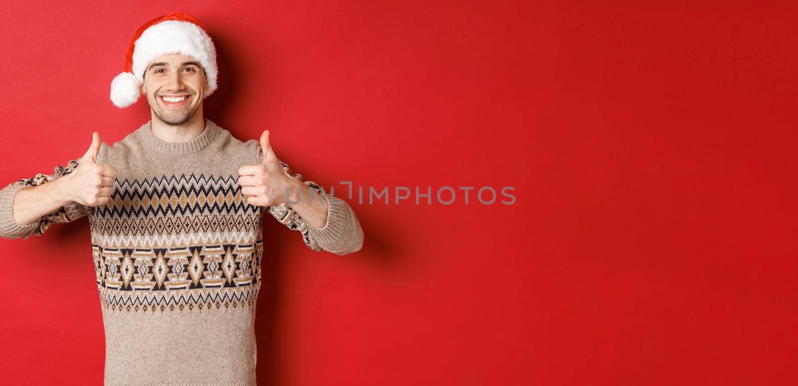 Image of cheerful attractive man in winter sweater and santa hat, showing thumbs-up, celebrating christmas and looking happy, like something, standing over red background.