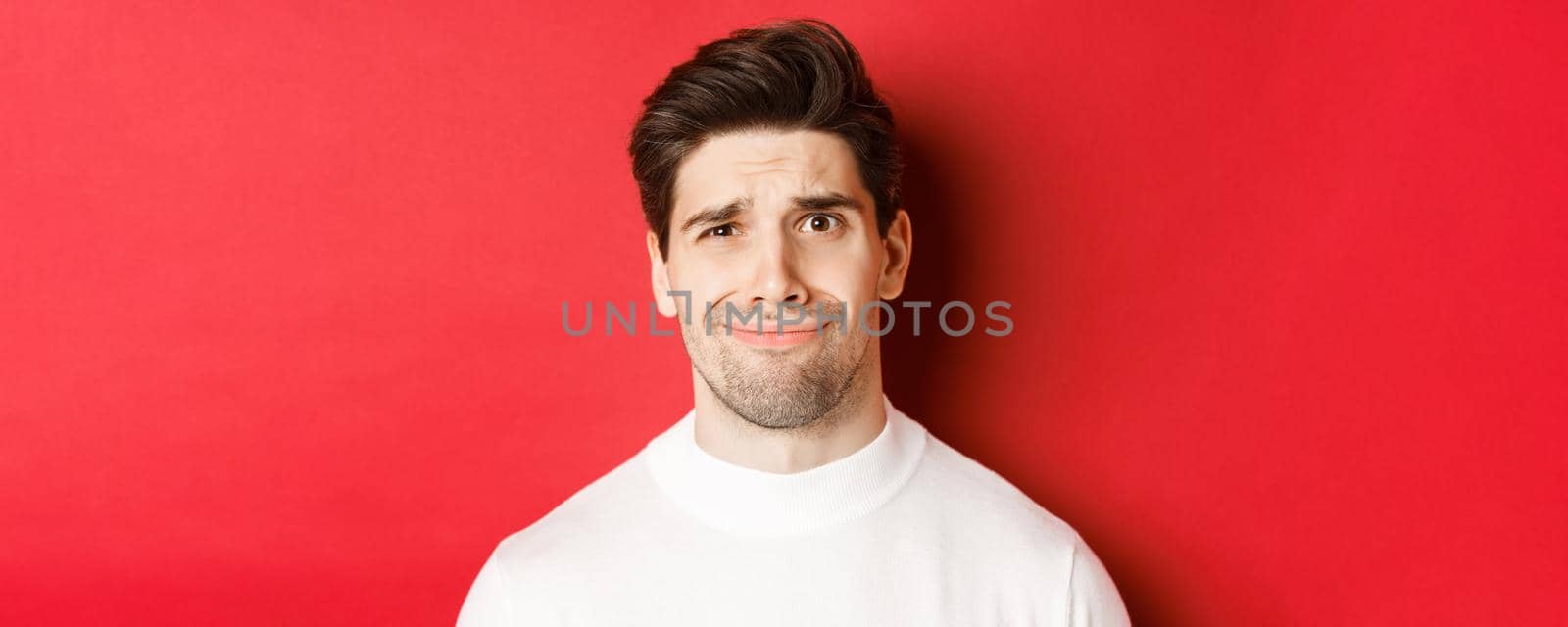 Close-up of handsome man feeling uncomfortable, grimacing and looking at something displeasing, standing over red background.