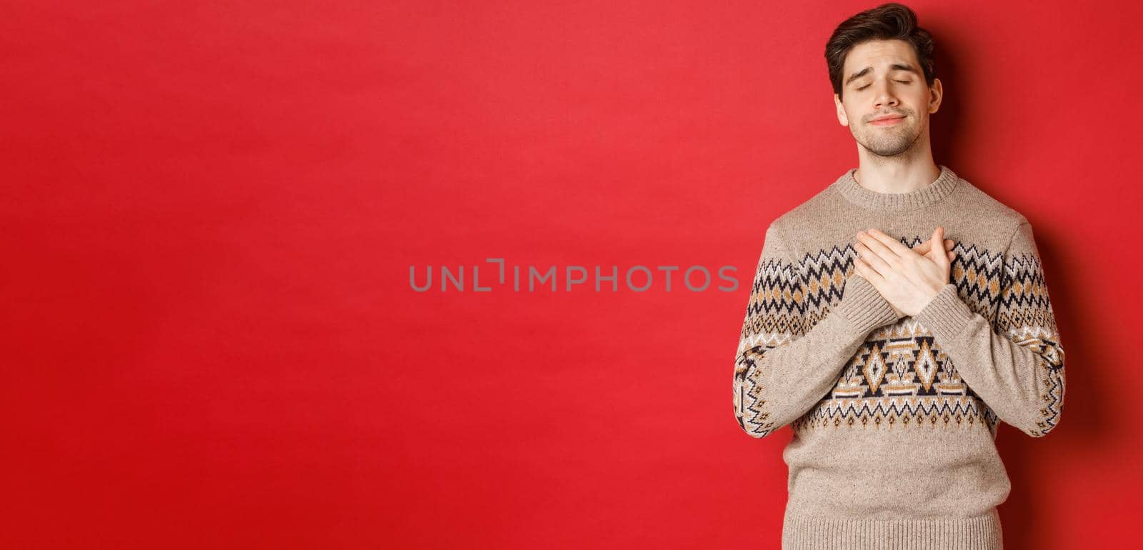 Image of dreamy and happy, attractive bearded man in christmas sweater, holding hands on heart, smiling with eyes closed, remember something touching, celebrating new year, red background by Benzoix