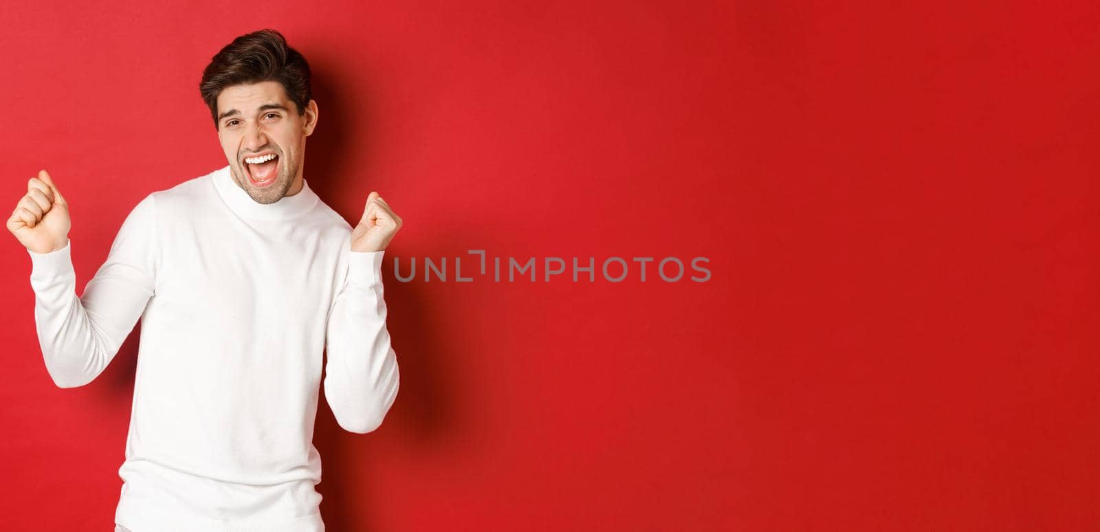 Portrait of handsome man enjoying new year party, dancing and having fun, standing in white sweater against red background. Concept of winter holidays and christmas.