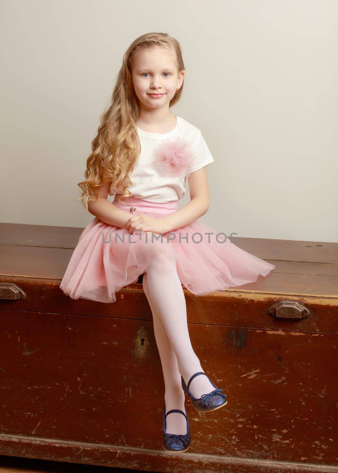 Beautiful little blonde girl with long hair.She sits on an old trunk with her foot on her leg.In a lush short pink skirt and white T-shirt.