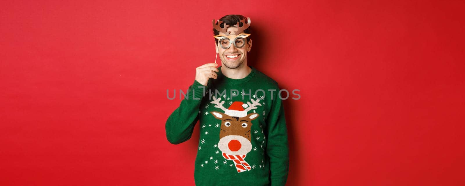 Handsome happy man in christmas sweater, holding party mask and smiling, enjoying new year celebration, standing over red background.