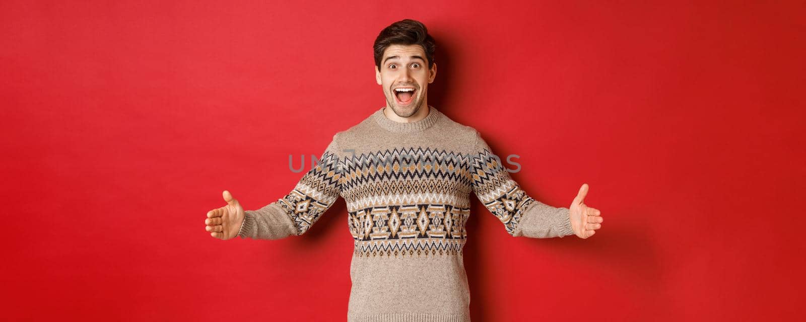 Portrait of attractive happy man in christmas sweater showing big present, spread hands sideways as if holding large box, standing over red background.