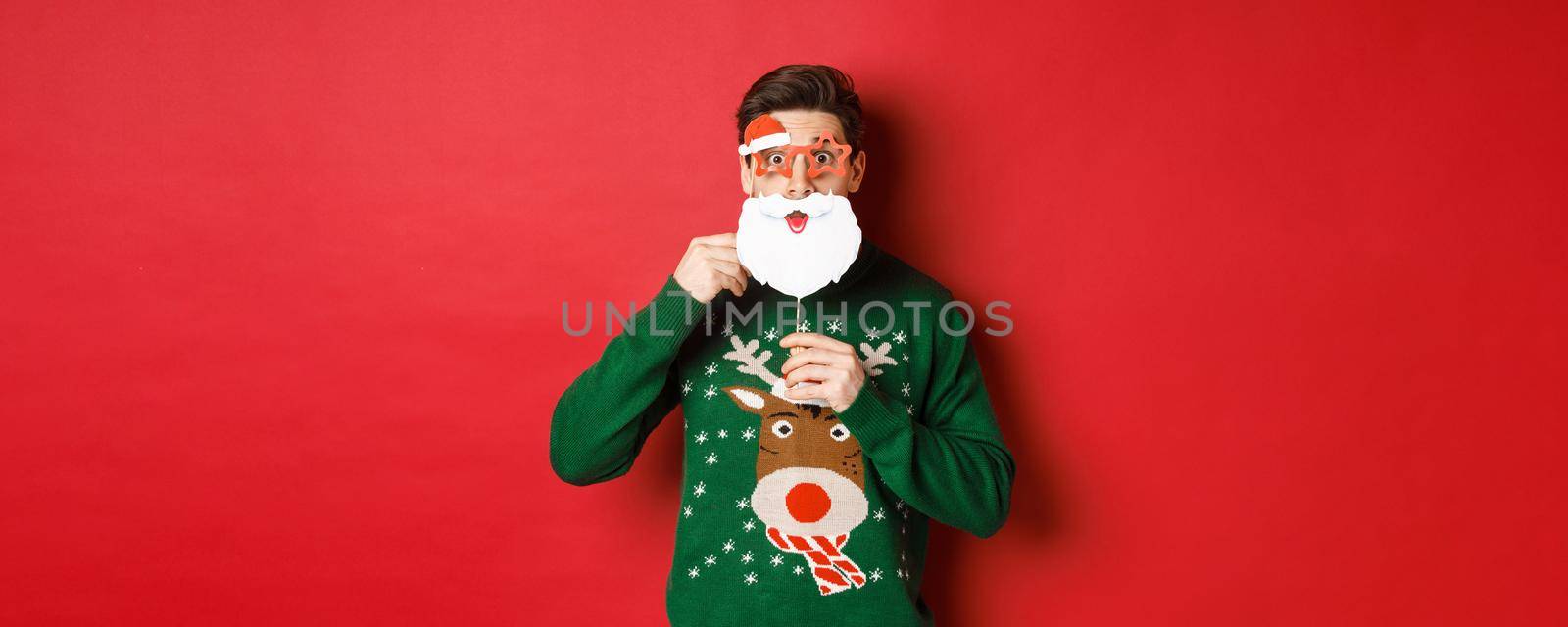 Portrait of surprised man in green christmas sweater, holding funny santa claus mask, celebrating new year, standing over red background.