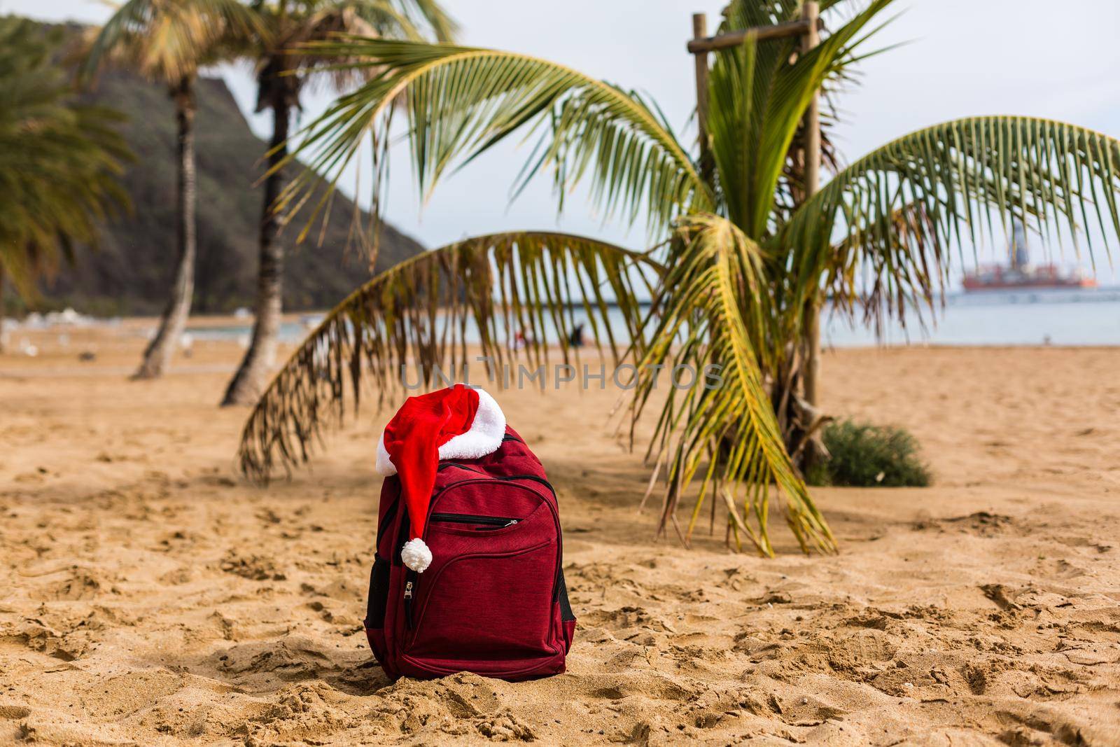 Santa hat on a tourist red backpack against the blue sea, enjoying the sunset of the ocean horizon. Christmas beach vacation travel banner or card