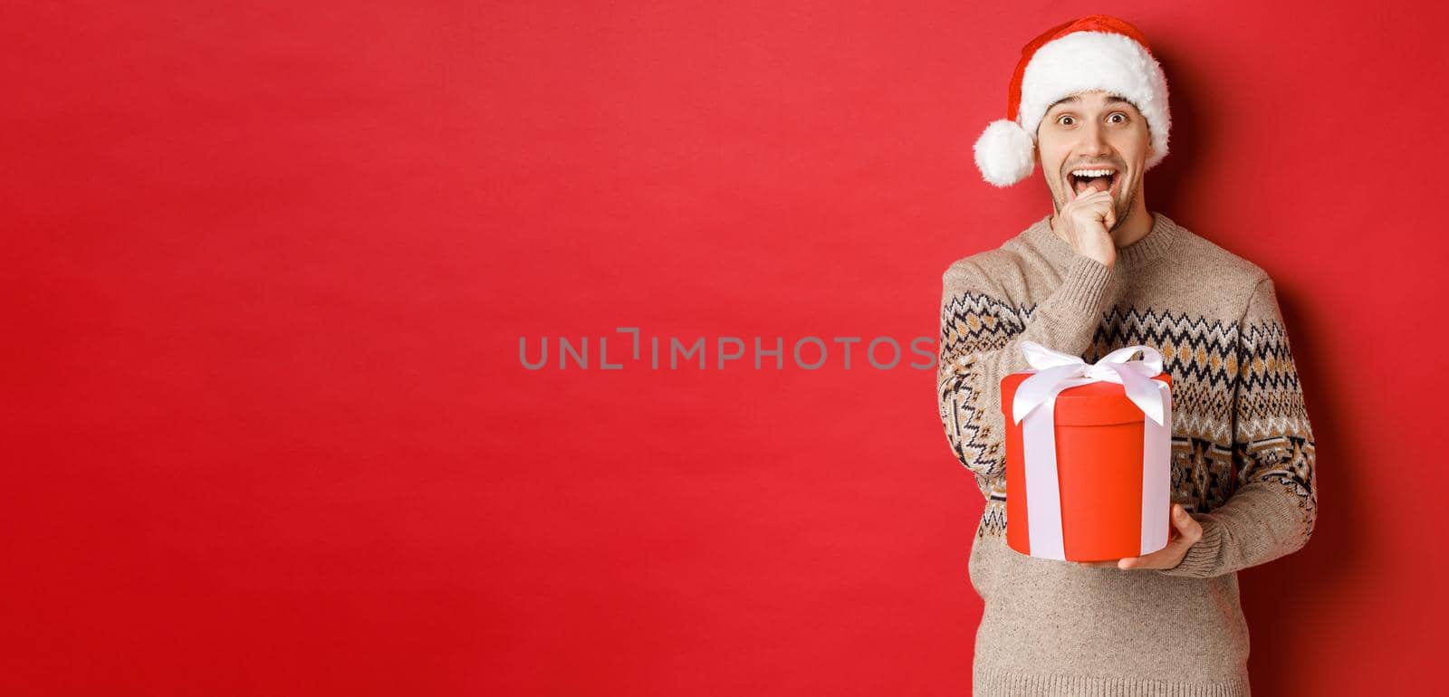 Image of handsome guy in santa hat and christmas sweater, excited to open xmas gift, looking amazed and holding present in one hand, standing over red background.