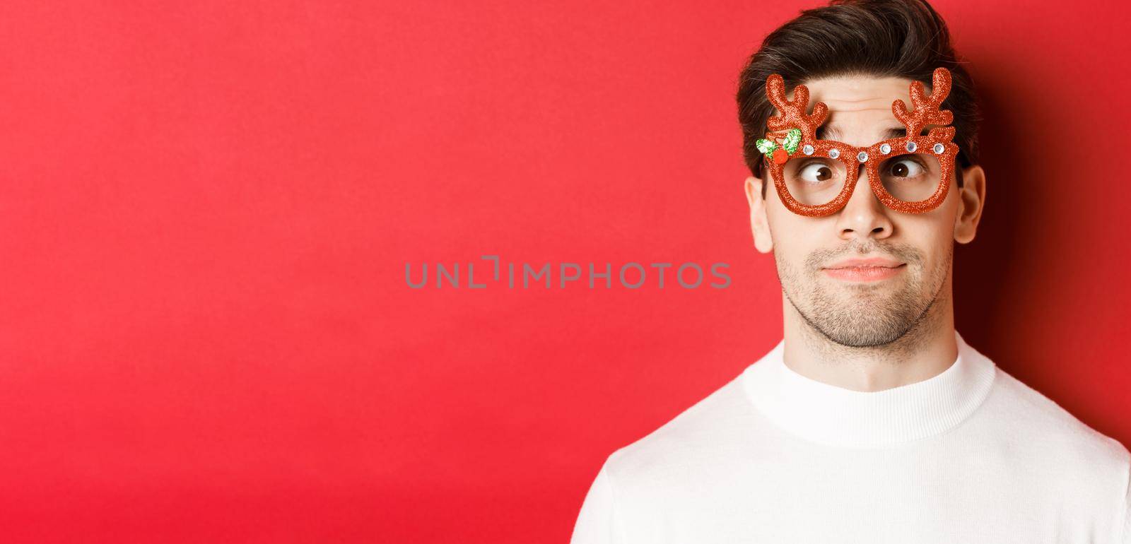 Concept of winter holidays, christmas and celebration. Close-up of funny brunette in party glasses, squinting and making faces, standing over red background.