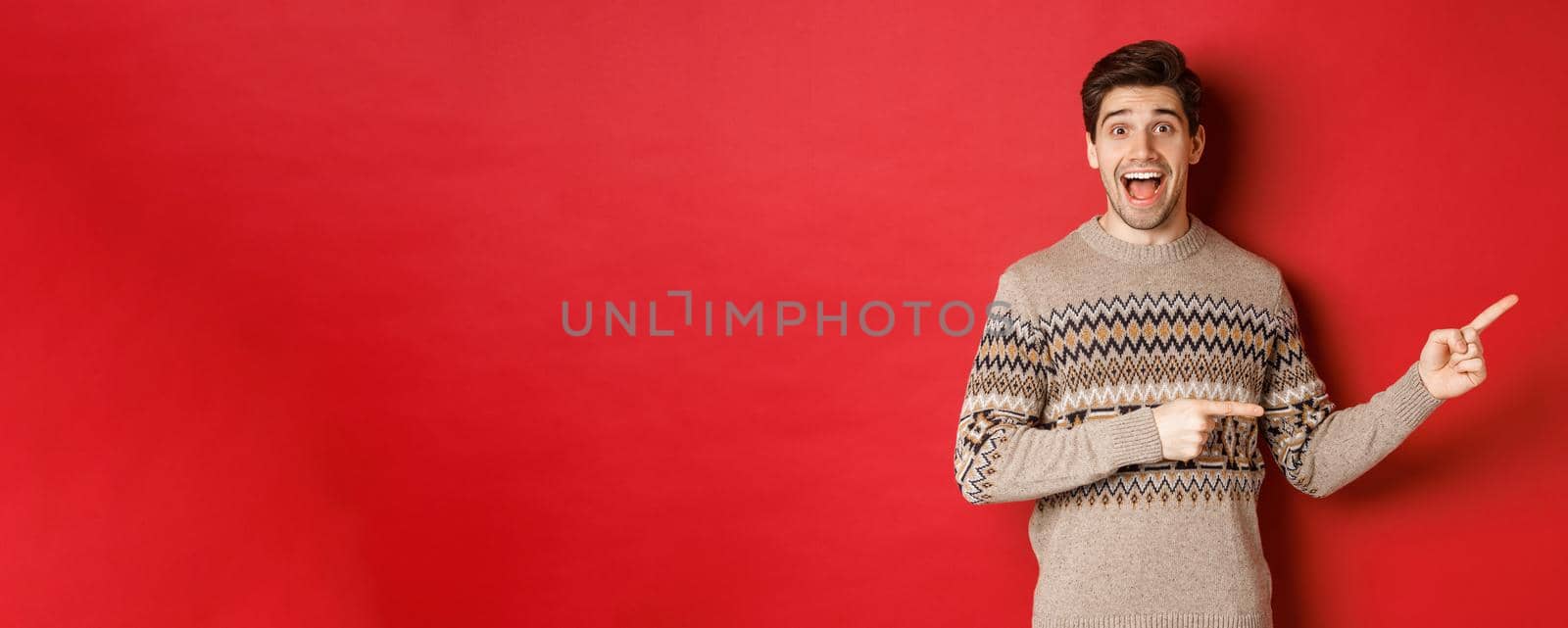 Concept of christmas celebration, winter holidays and lifestyle. Handsome man in xmas sweater pointing fingers right, smiling amazed, showing new year promo against red background by Benzoix