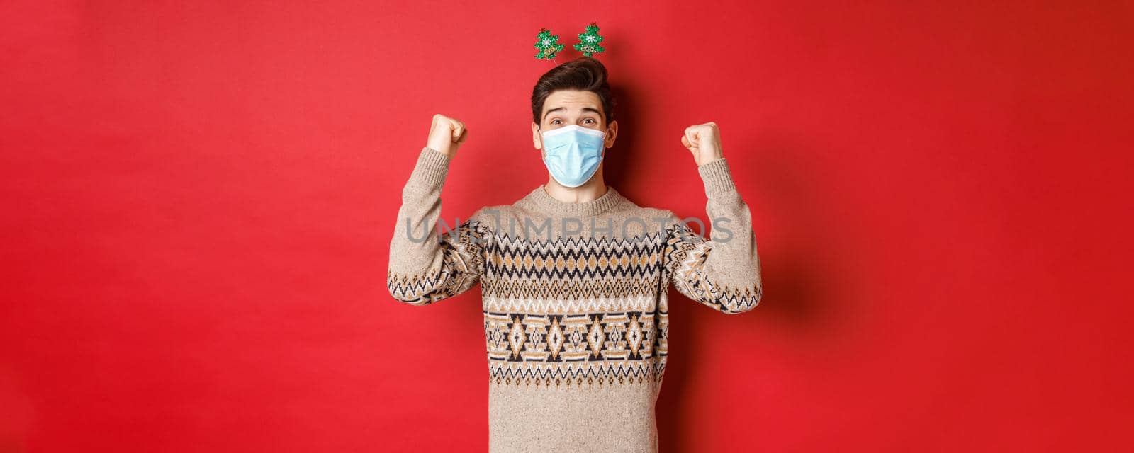 Concept of christmas, holidays and coronavirus. Happy man celebrating new year during covid-19 outbreak, wearing medical mask and sweater, rejoicing over red background by Benzoix