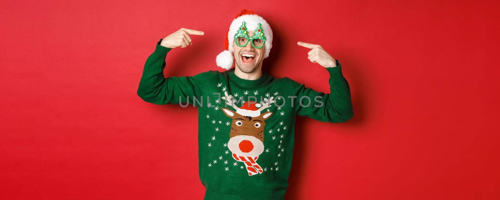 Image of excited smiling guy pointing at party glasses and celebrating new year, standing joyful in green sweater and santa hat against red background.