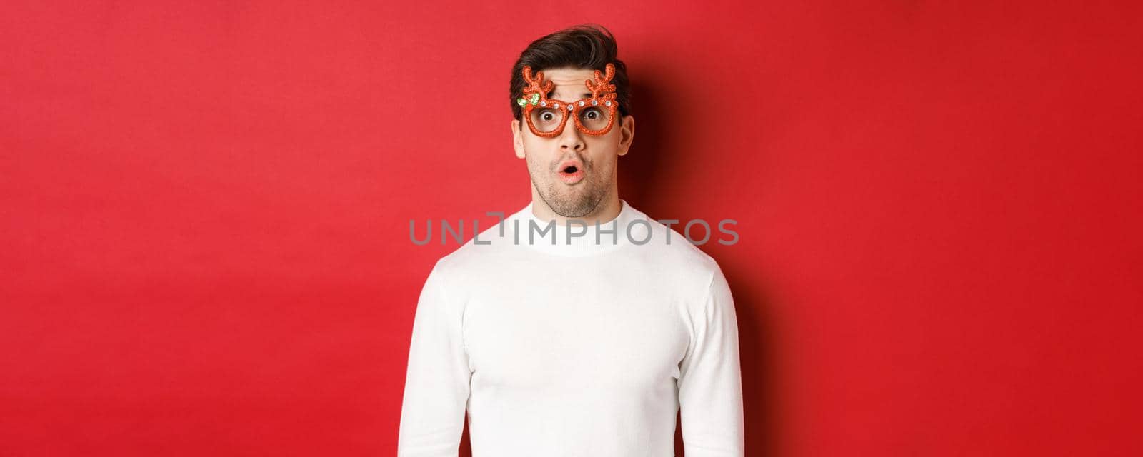 Close-up of surprised and impressed man in white sweater, party glasses, saying wow and looking amazed at camera, standing over red background by Benzoix