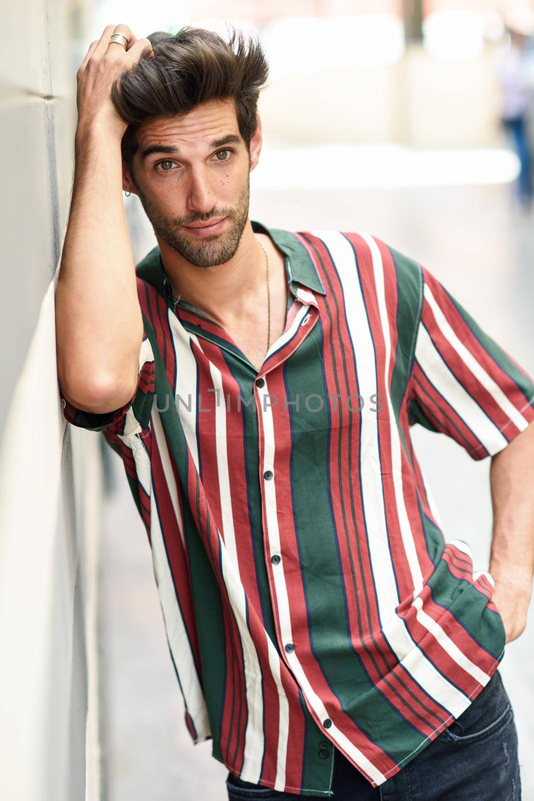 Attractive young man with dark hair and modern hairstyle wearing casual clothes in urban background.
