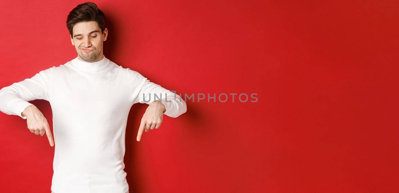 Portrait of unamused handsome man, looking skeptical while pointing fingers down at something bad, standing over red background.