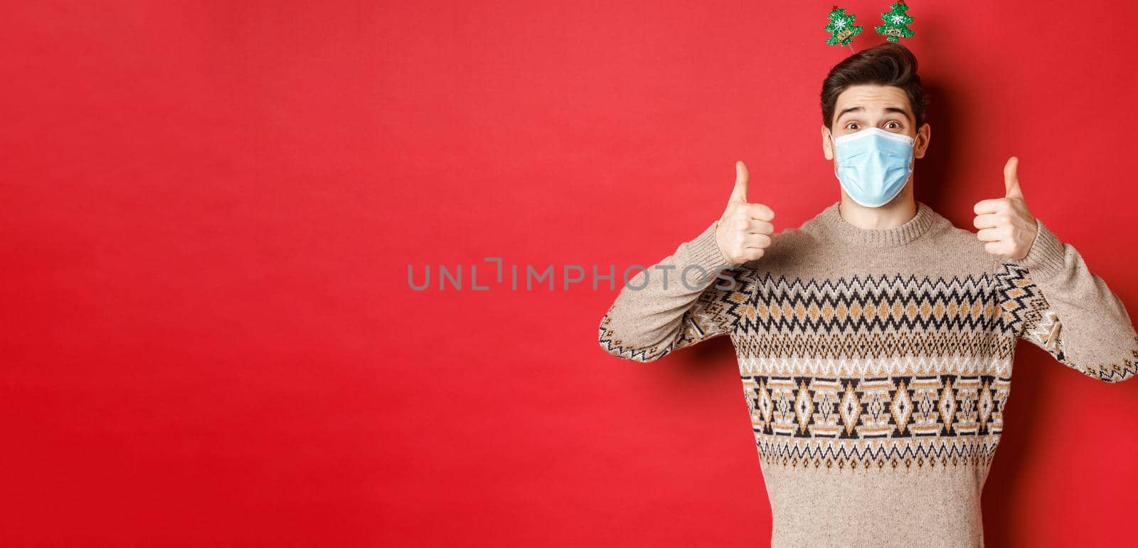 Concept of new year, covid-19 and social distancing. Cheerful caucasian man in sweater and medical mask, celebrating christmas during pandemic, showing thumbs-up, red background.