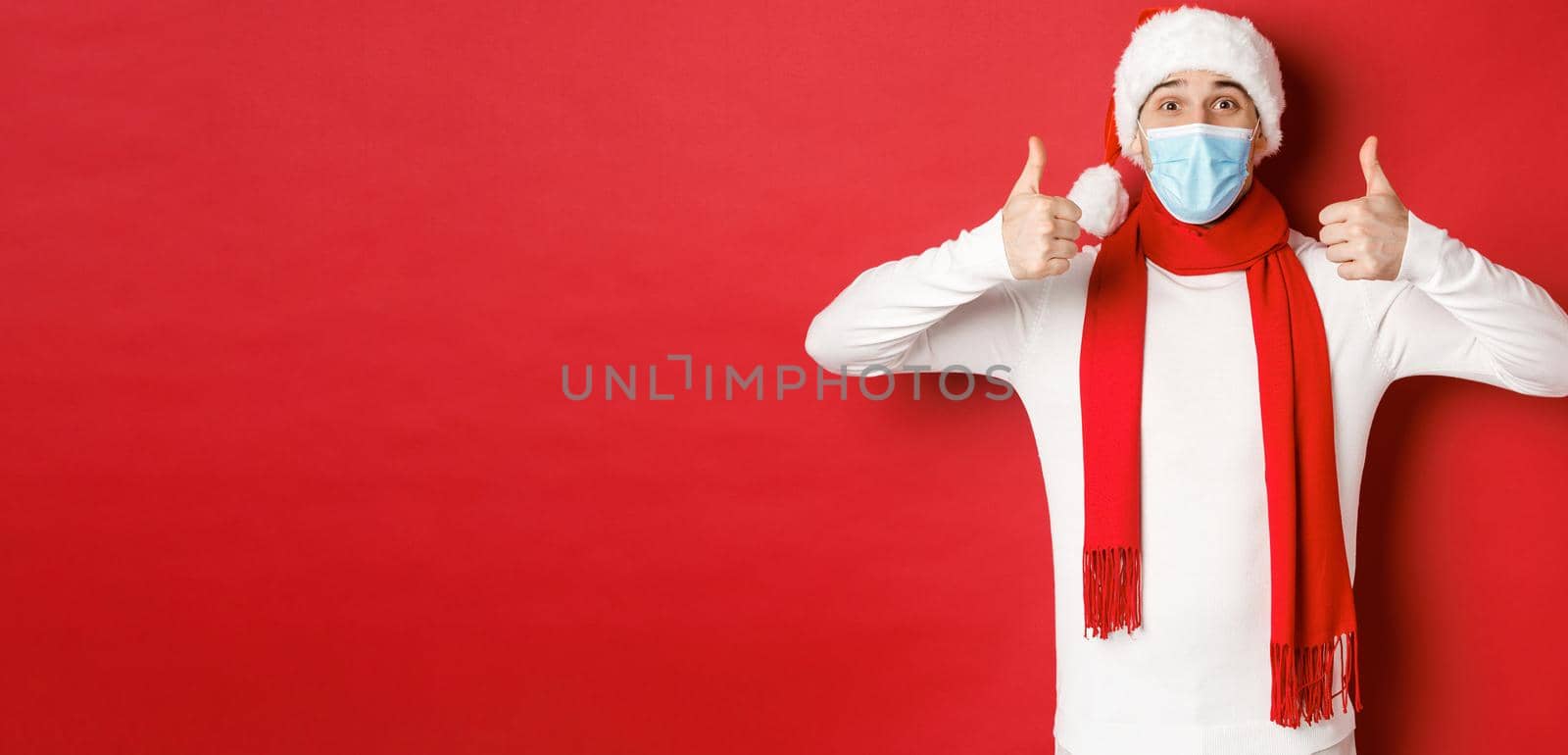 Concept of new year, coronavirus and holidays. Cheerful man celebrating new year and social distancing, wearing medical mask, santa hat and scarf, showing thumbs-up in approval.