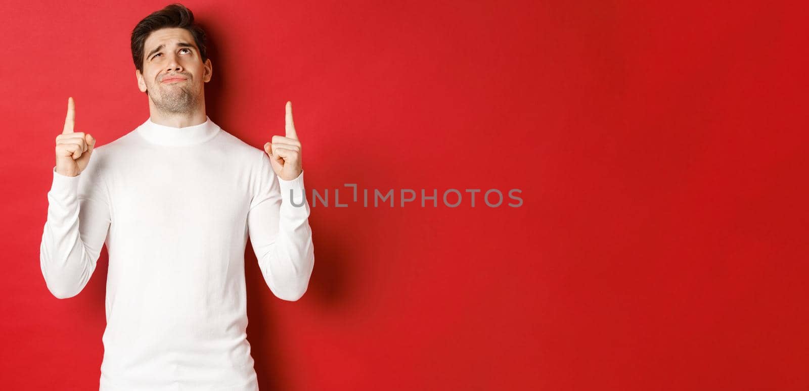 Concept of winter holidays. Doubtful handsome man in white sweater, looking skeptical and pointing fingers up at logo, standing against red background.