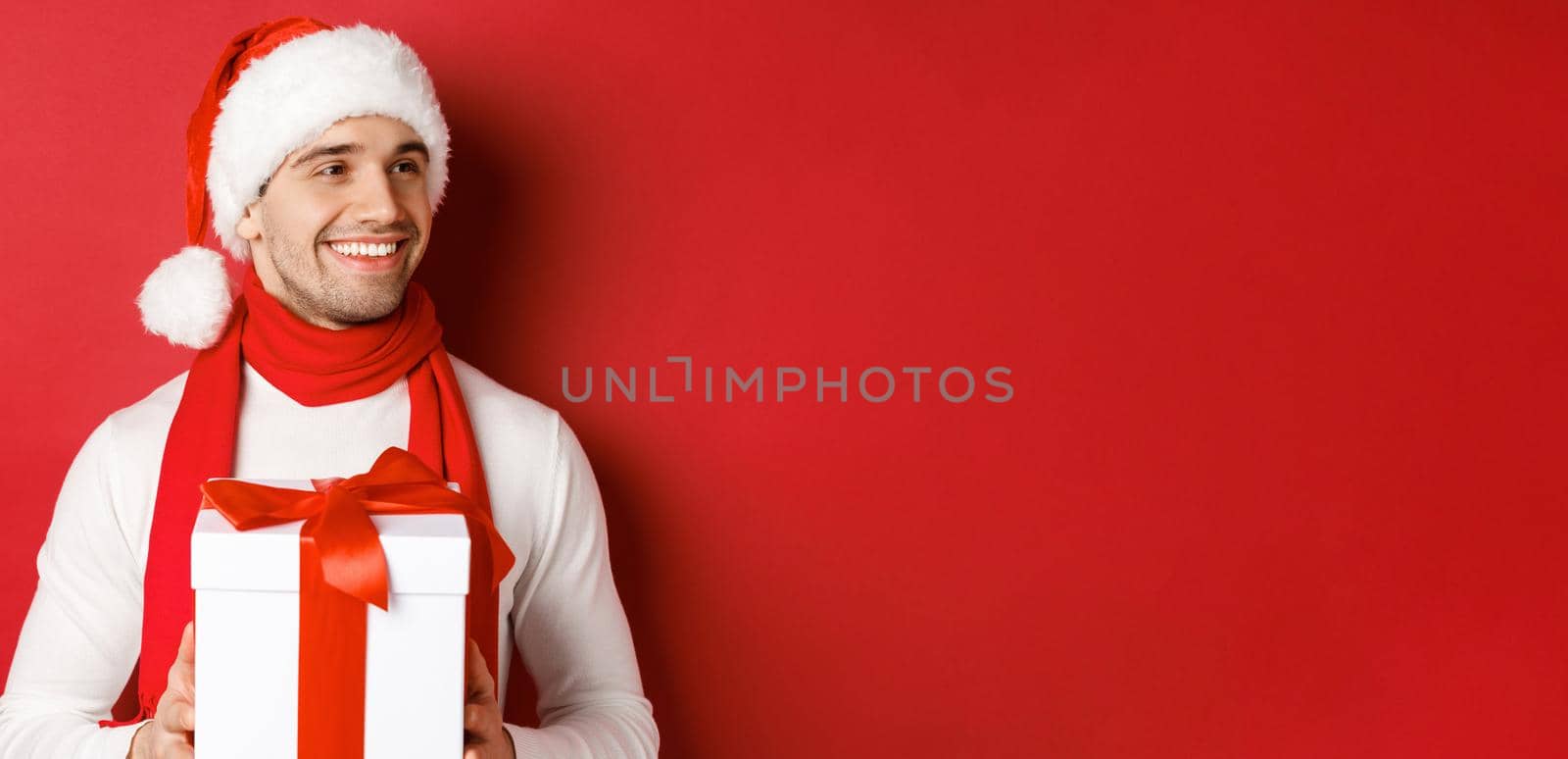 Concept of winter holidays, christmas and lifestyle. Close-up of attractive man in santa hat and scarf, holding new year gift, looking right and smiling, standing over red background.