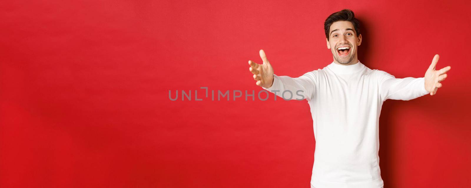 Image of happy, friendly man in white sweater, reaching hands and smiling while greeting guests to christmas party, waiting for hug, standing over red background.