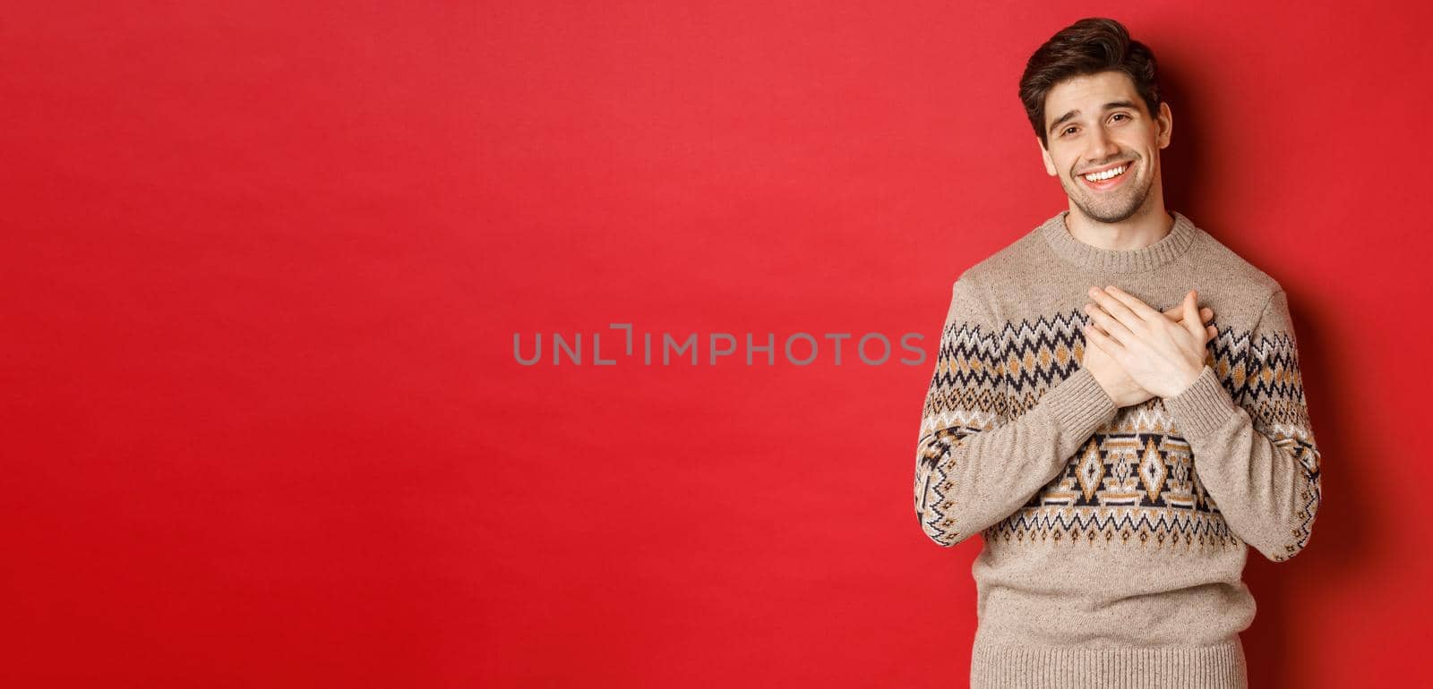 Portrait of touched and happy handsome guy, receiving new year gift, holding hands on heart and smiling, saying thank you, standing in christmas sweater against red background.