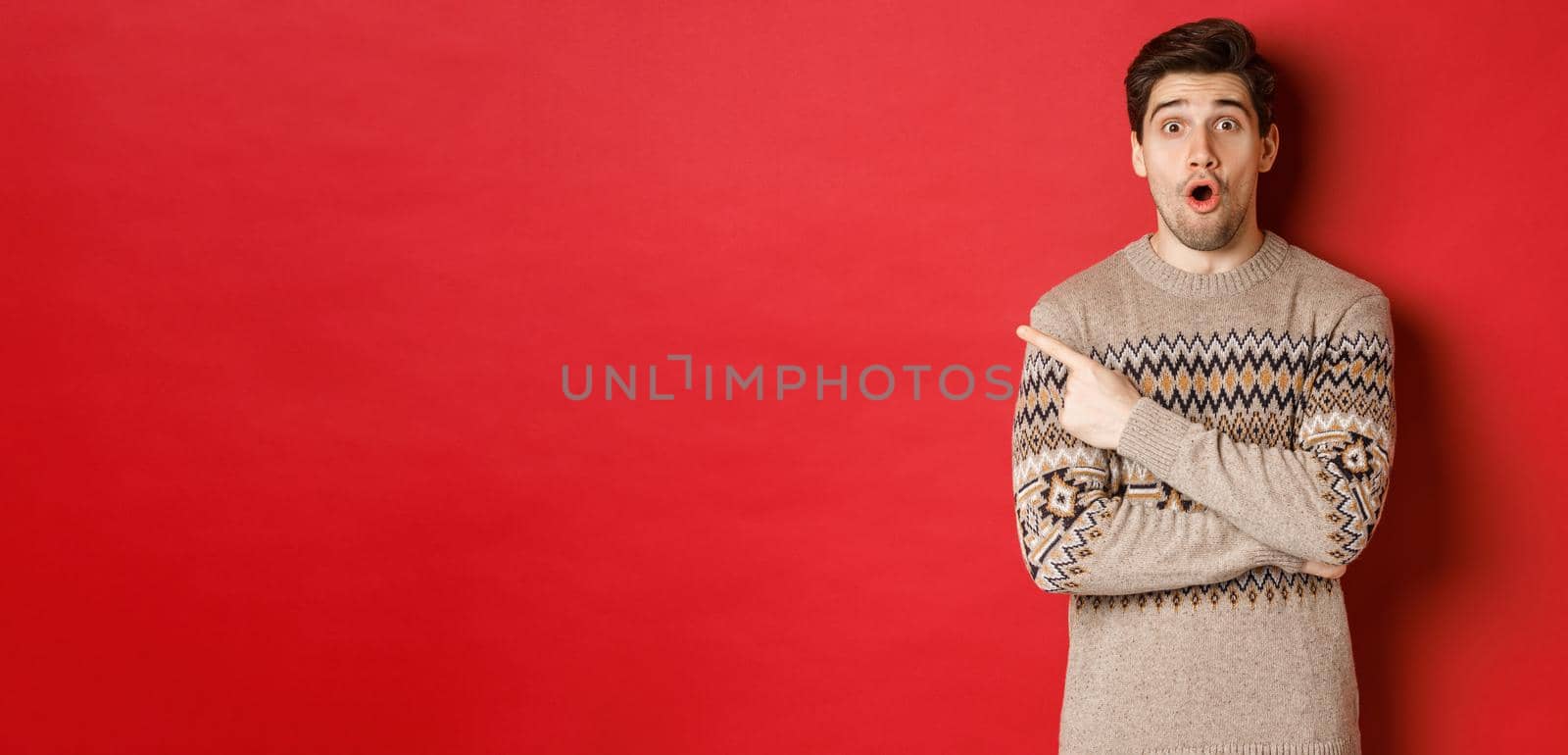 Image of impressed handsome guy, showing awesome christmas promo offer, pointing finger at upper left corner and looking amazed, standing in xmas sweater over red background.