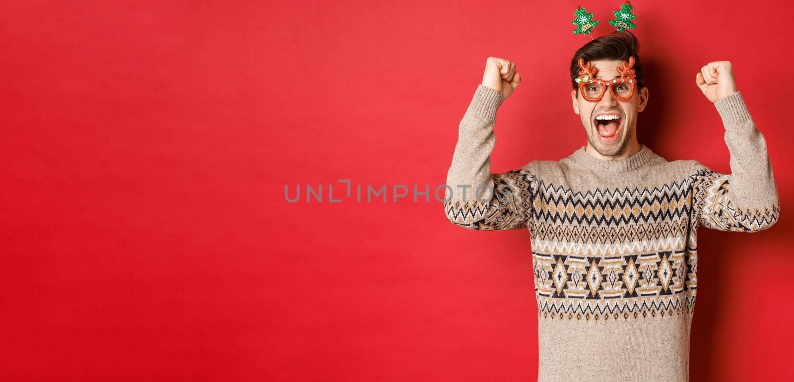 Portrait of thrilled and happy man in party glasses, winter sweater, shouting for joy and raising hands up, winning prize on christmas, achieve new year goal, standing red background.