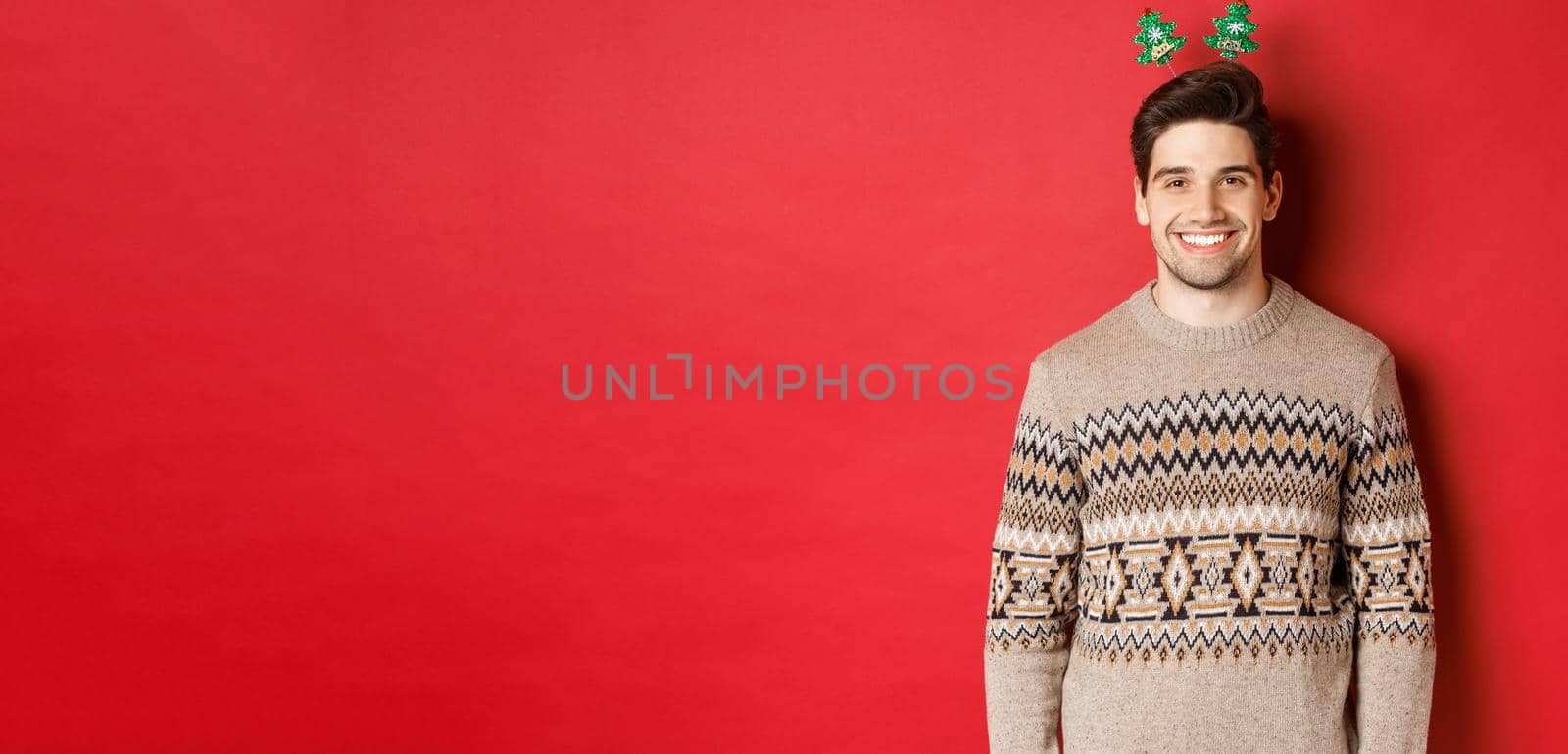 Concept of winter holidays, christmas and celebration. Handsome bearded guy in sweater wishing happy new year, smiling at camera, standing over red background by Benzoix