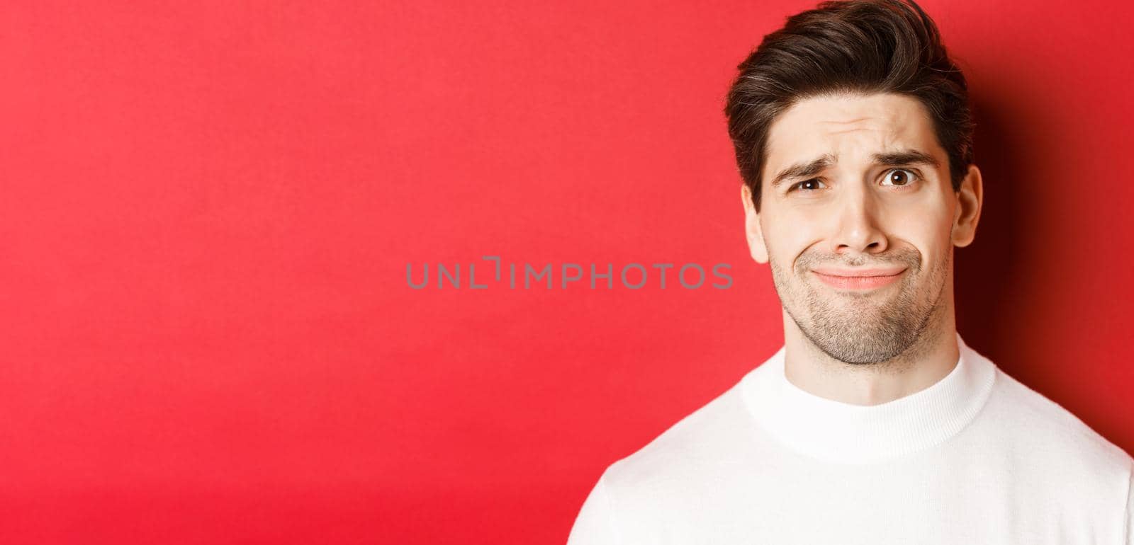 Close-up of handsome man feeling uncomfortable, grimacing and looking at something displeasing, standing over red background by Benzoix