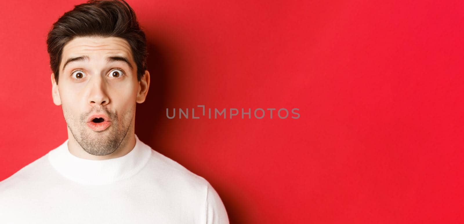 Close-up of surprised handsome man in white sweater, open mouth and looking wondered, hear amazing news, standing over red background.