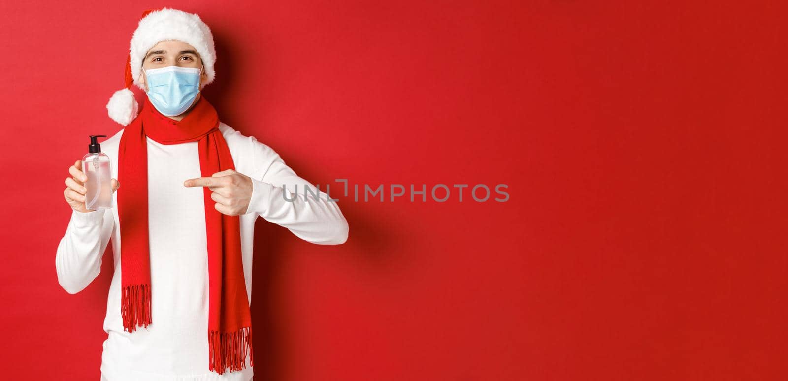 Concept of covid-19, christmas and holidays during pandemic. Handsome happy man in santa hat and medical mask, recommending use hand sanitizer, standing over red background.