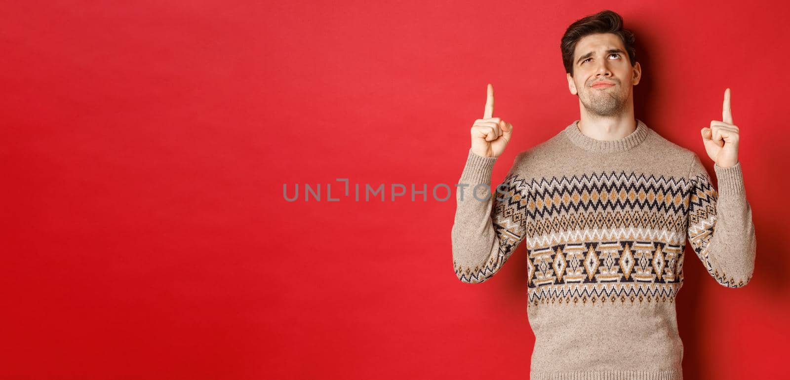 Image of disappointed and skeptical handsome man, wearing christmas sweater, smirk and frowning as looking at something bad, pointing fingers up, standing over red background by Benzoix