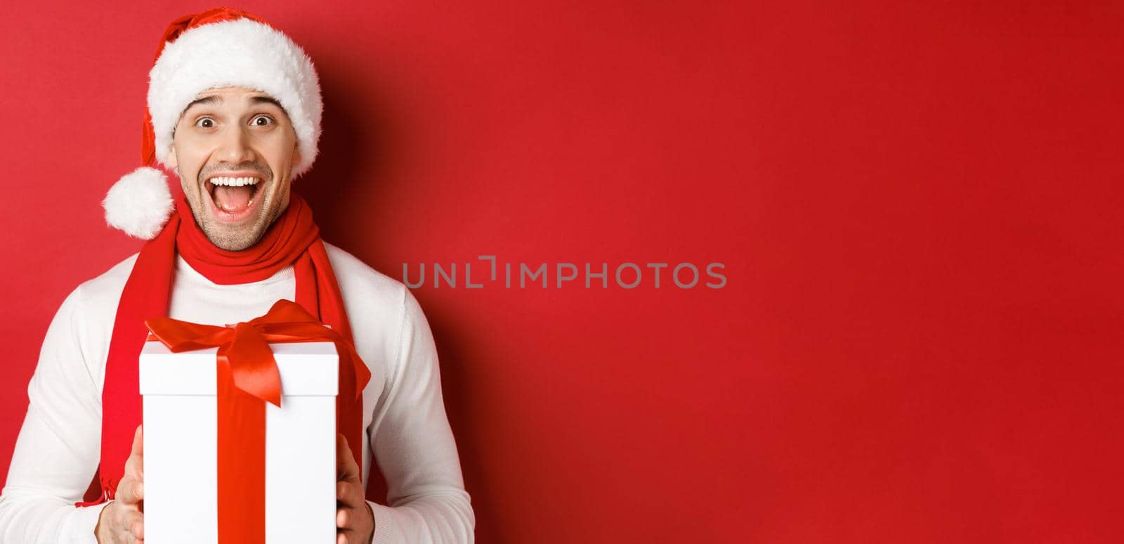 Concept of winter holidays, christmas and lifestyle. Close-up of excited handsome man in santa jat and scarf, looking amazed and receiving new year present, standing over red background.