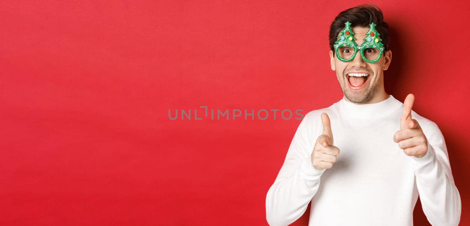 Joyful caucasian guy in party glasses and white sweater, smiling and pointing fingers at camera, wishing merry christmas and happy new year, standing over red background.