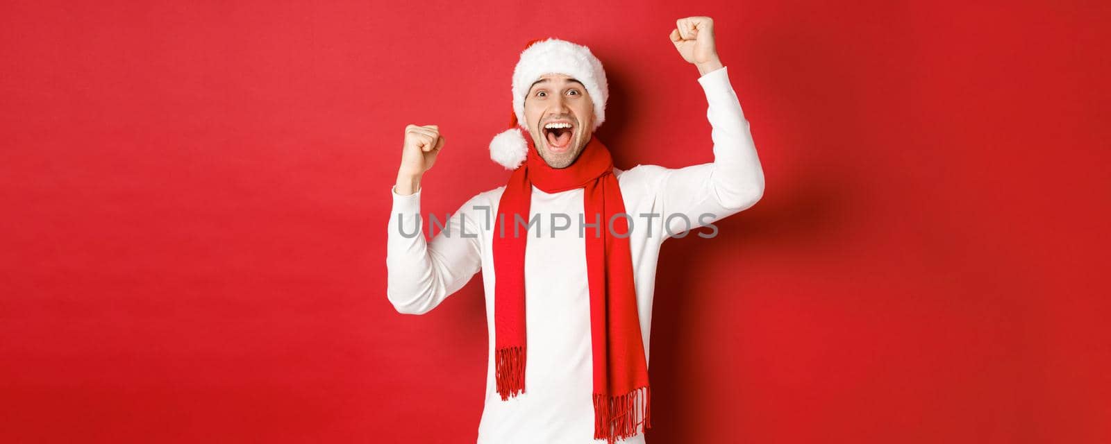 Image of joyful caucasian man in santa hat and scarf, shouting for joy and raising hands, celebrating victory or win, triumphing over red background by Benzoix