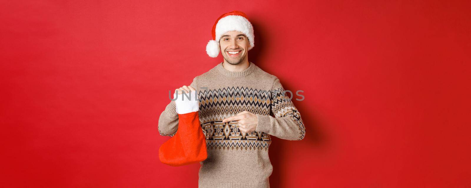 Concept of winter holidays, new year and celebration. Handsome smiling man prepared gifts for kids, pointing at christmas stocking bag, standing over red background by Benzoix