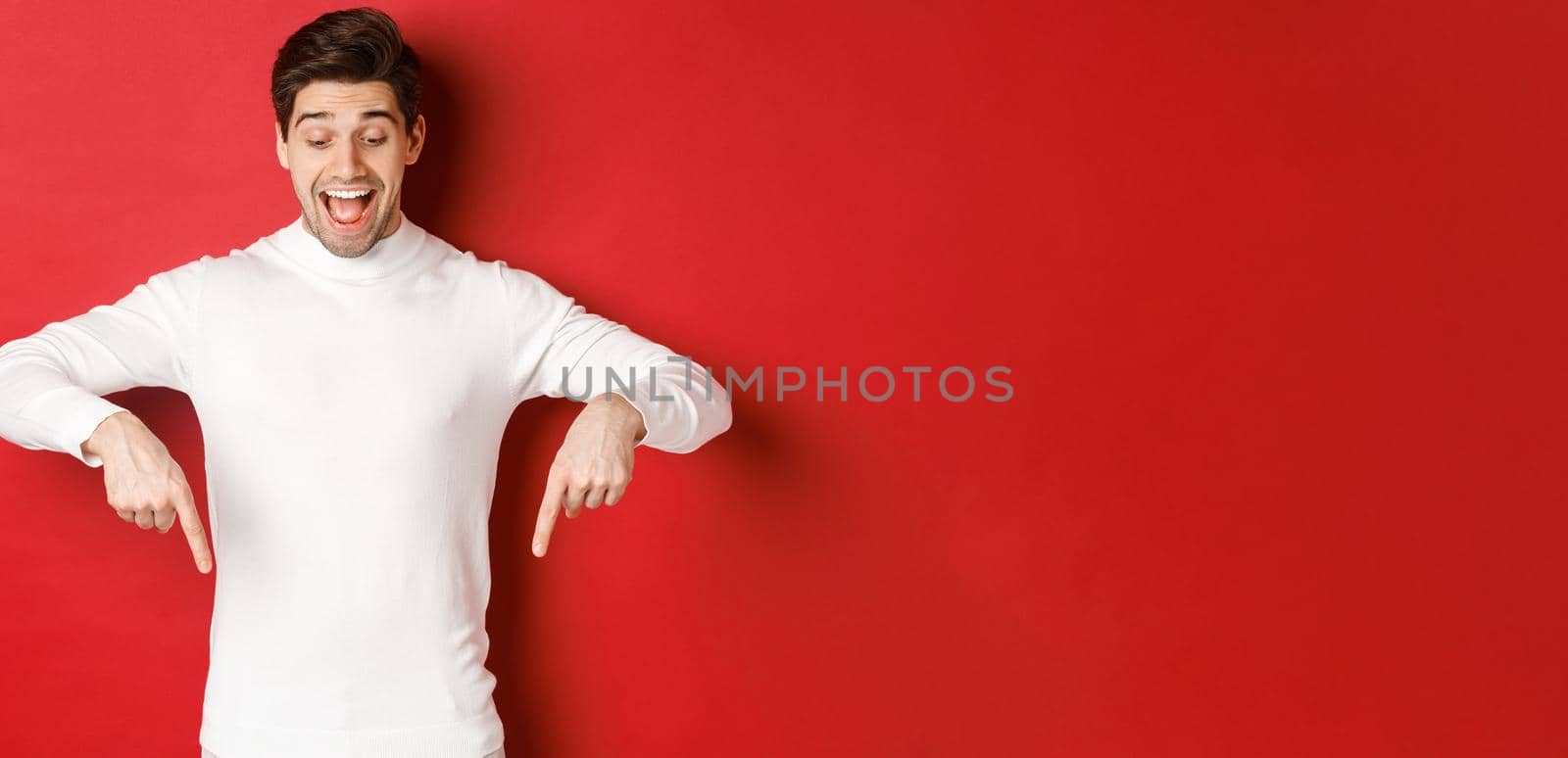 Portrait of happy good-looking man, pointing fingers down and looking at advertisement with excited smile, standing over red background.